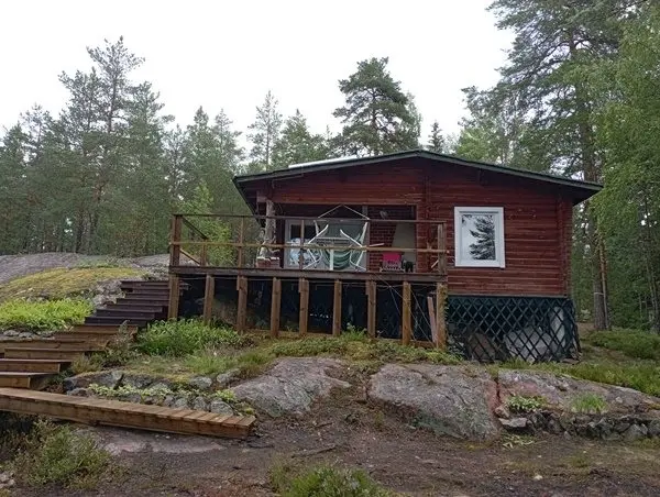 Tumma hirsimökki kalliolla ennen peruskorjausta, vanha terassi ja portaat männikössä